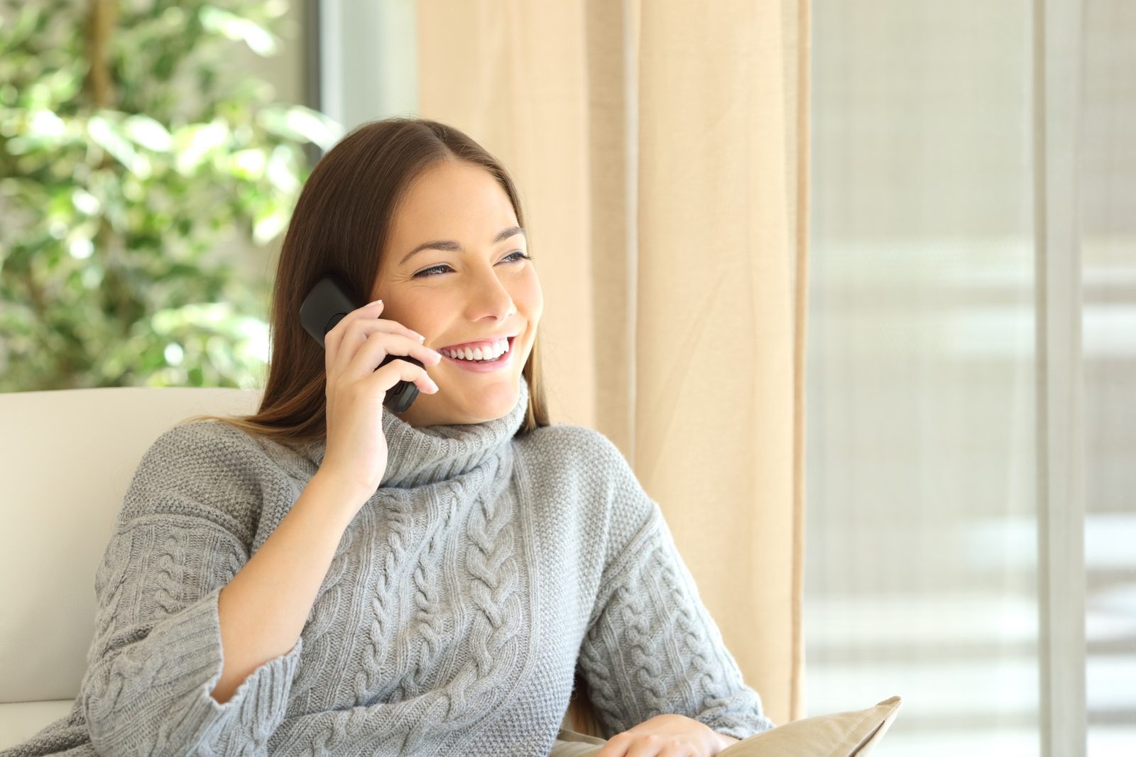 A woman in a gray sweater laughing on a cordless home phone on the couch